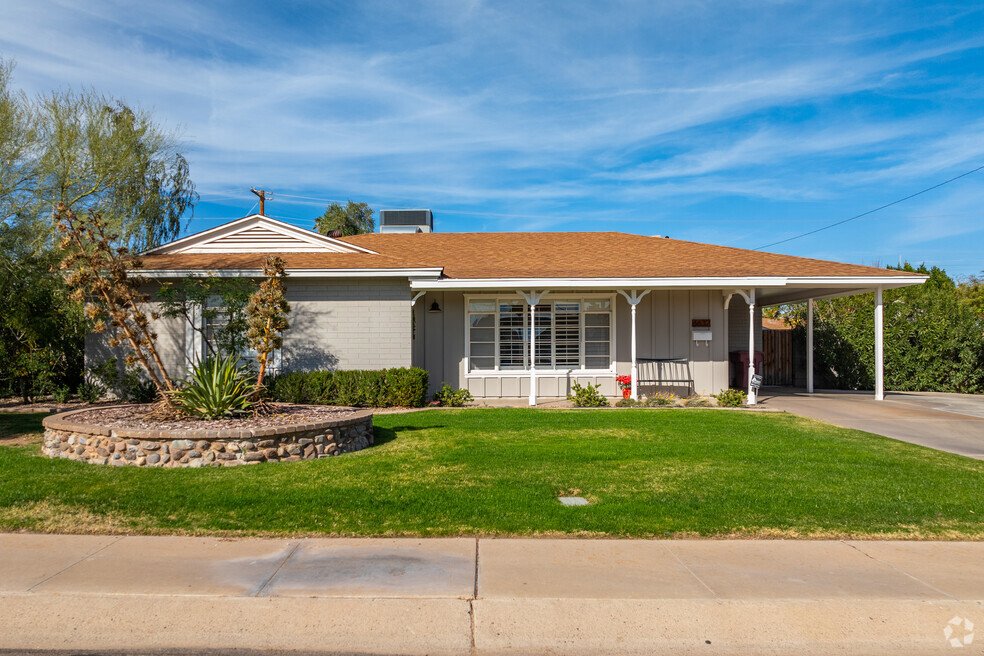 The house is primarily light gray or beige brick, with a brown tiled roof that extends to create a covered porch running across the front of the house. White trim accents the windows and the porch's supporting beams, providing a crisp contrast to the brick. The porch features a simple, dark-colored bench. A carport is attached to the side of the house, providing covered parking.
