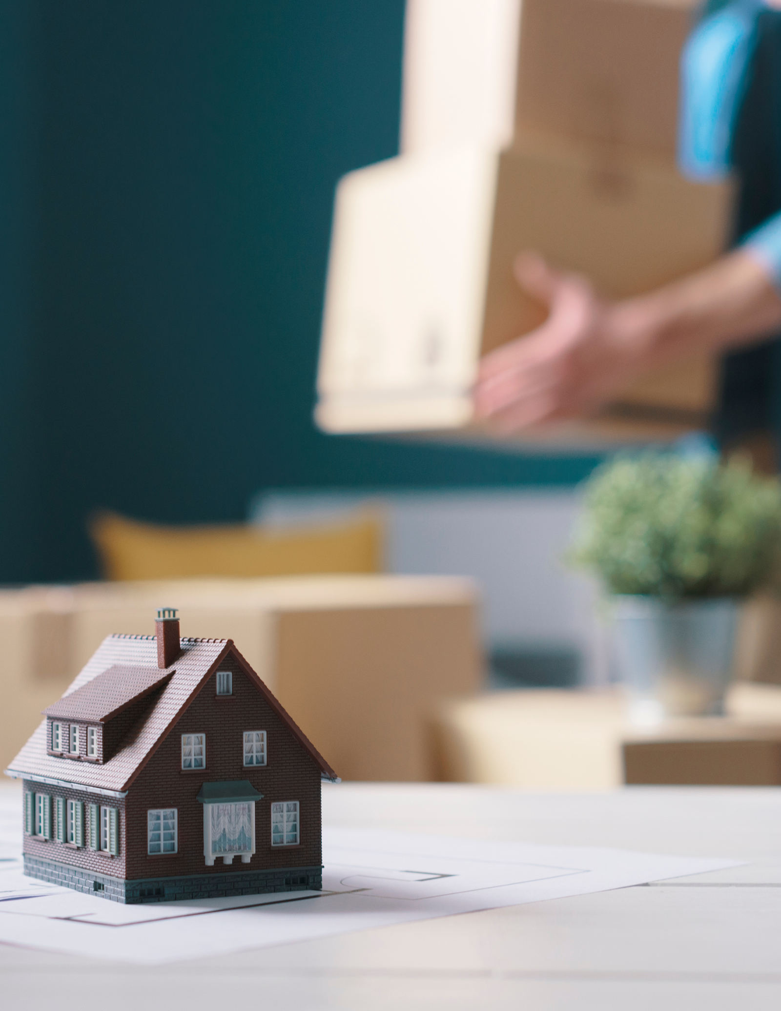 A small model house sits on a table in the foreground, with a blurred background showing a person carrying boxes and other moving supplies. The image evokes a sense of moving into a new house.