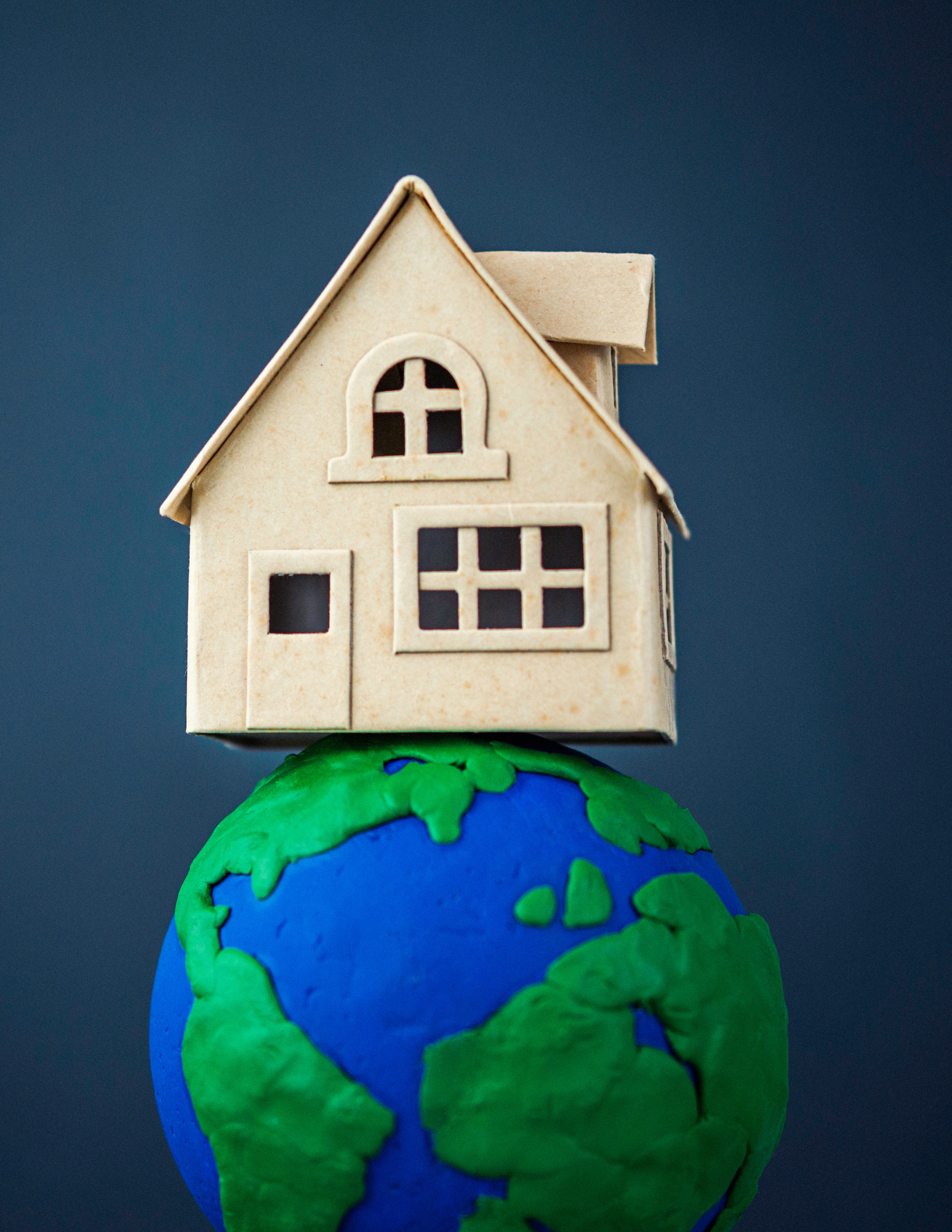 A small model house made of light brown cardboard sits atop a miniature globe crafted from blue and green clay, set against a dark blue background. The image symbolizes the global nature of housing or real estate.